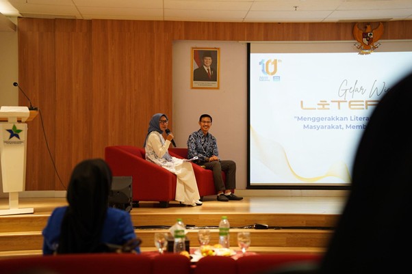 gelar wicara 10th sli 2 dompet dhuafa Two speakers seated on a red couch on a stage, presenting to an audience with a large screen beside them.