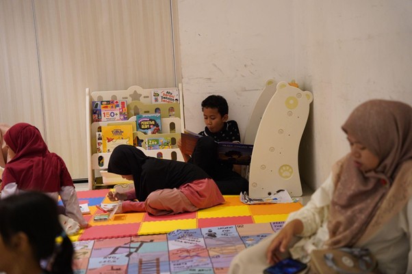gelar wicara 10th sli 6 dompet dhuafa Children’s reading corner with colorful foam mats, a bookshelf, and kids reading books; two women in hijabs sit nearby.