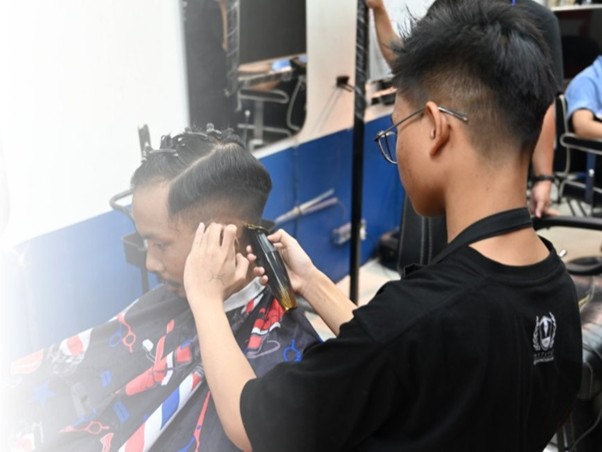 industribarberinstitutkemandirian3 dompet dhuafa Barber trims a man's hair with clippers while he sits in a barber chair with a cape draped over him in a barbershop.