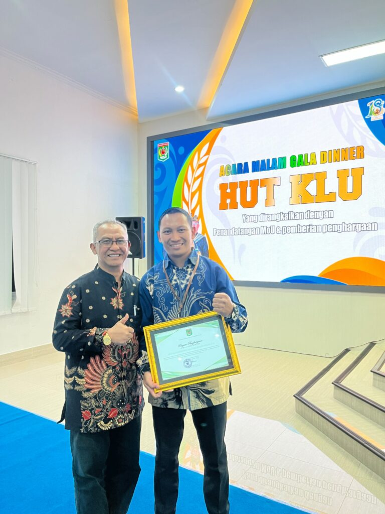 lkc dd ntb terima penghargaan 3 dompet dhuafa Two men in patterned shirts pose for a photo at an award event, one holding a framed certificate in front of a colorful banner screen.