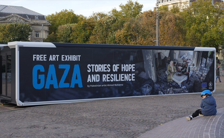 lukisansenimangazapalestina2768x473 dompet dhuafa Mobile art exhibit trailer with a banner: 'Free Art Exhibit Gaza – Stories of Hope and Resilience'; a person sits on the curb nearby.