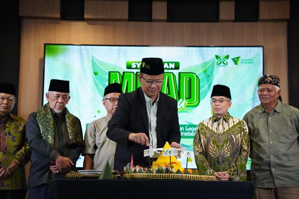 Man in a suit and black hat cuts a cake on a table, flanked by six men in traditional Indonesian attire at a stage with a green event banner.