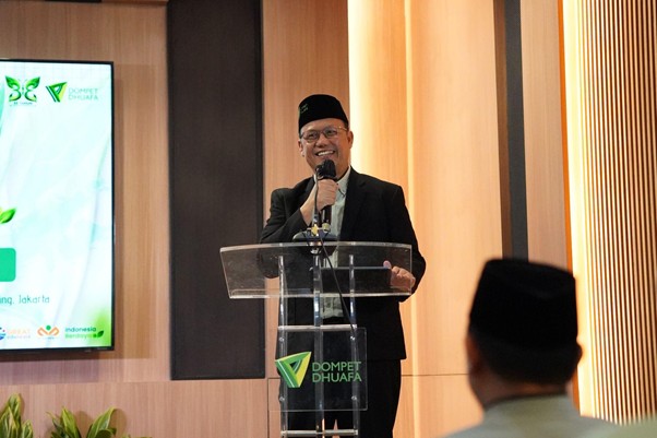 milad 33 th dompet dhuafa 2 dompet dhuafa A man in a suit and peci speaks at a clear podium with a microphone, in a conference room with a large screen beside him showing event graphics.