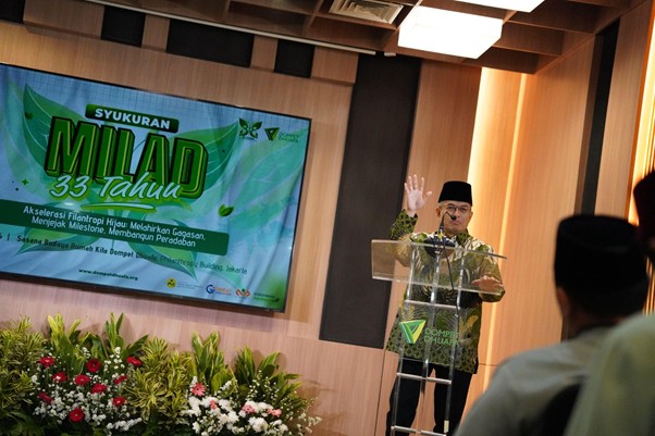 milad 33 th dompet dhuafa 3 dompet dhuafa Man in a black cap giving a speech at a podium on a decorated stage, with a banner reading 'MILAD 33 Tahun' and floral arrangements in front.