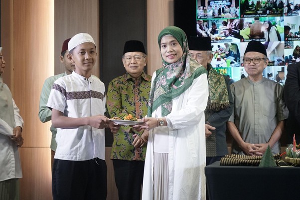 milad 33 th dompet dhuafa 6 dompet dhuafa A woman in a white dress with a green headscarf hands a plate to a young boy during a formal ceremony, with officials watching in the background.
