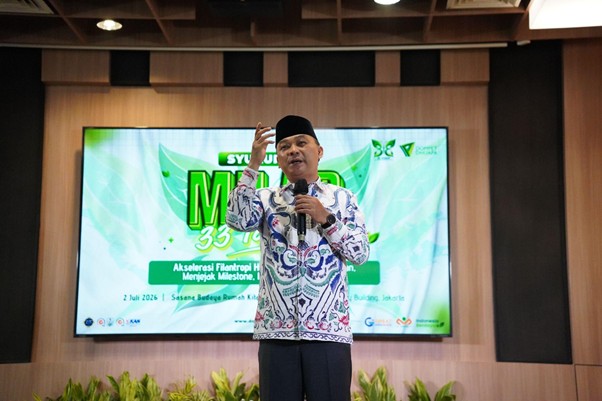 milad 33 th dompet dhuafa 7 dompet dhuafa Male speaker in batik and cap delivering a presentation on stage with a microphone, in front of a green event banner.