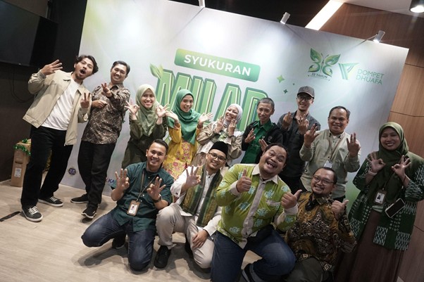 milad 33 th dompet dhuafa 9 dompet dhuafa Fifteen people posing together on a stage in a celebration event, smiling and showing peace or victory gestures, with a banner behind them reading 'Syukuran' and logos for the event.