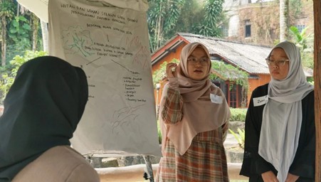 ngobrolbumi20263 dompet dhuafa Three women wearing hijabs stand around a large flip chart with a hand-drawn map, discussing outdoors amid tropical trees.