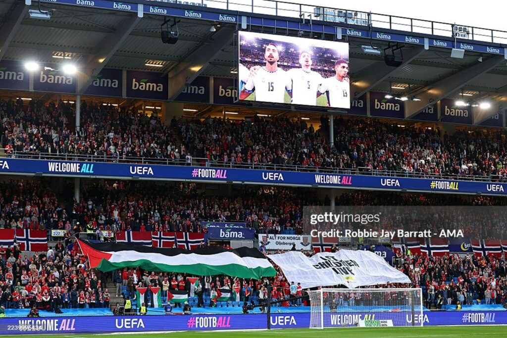 Packed football stadium crowd in red, waving flags under bright stadium lights; banners read 'WELCOME ALL' and 'UEFA'.