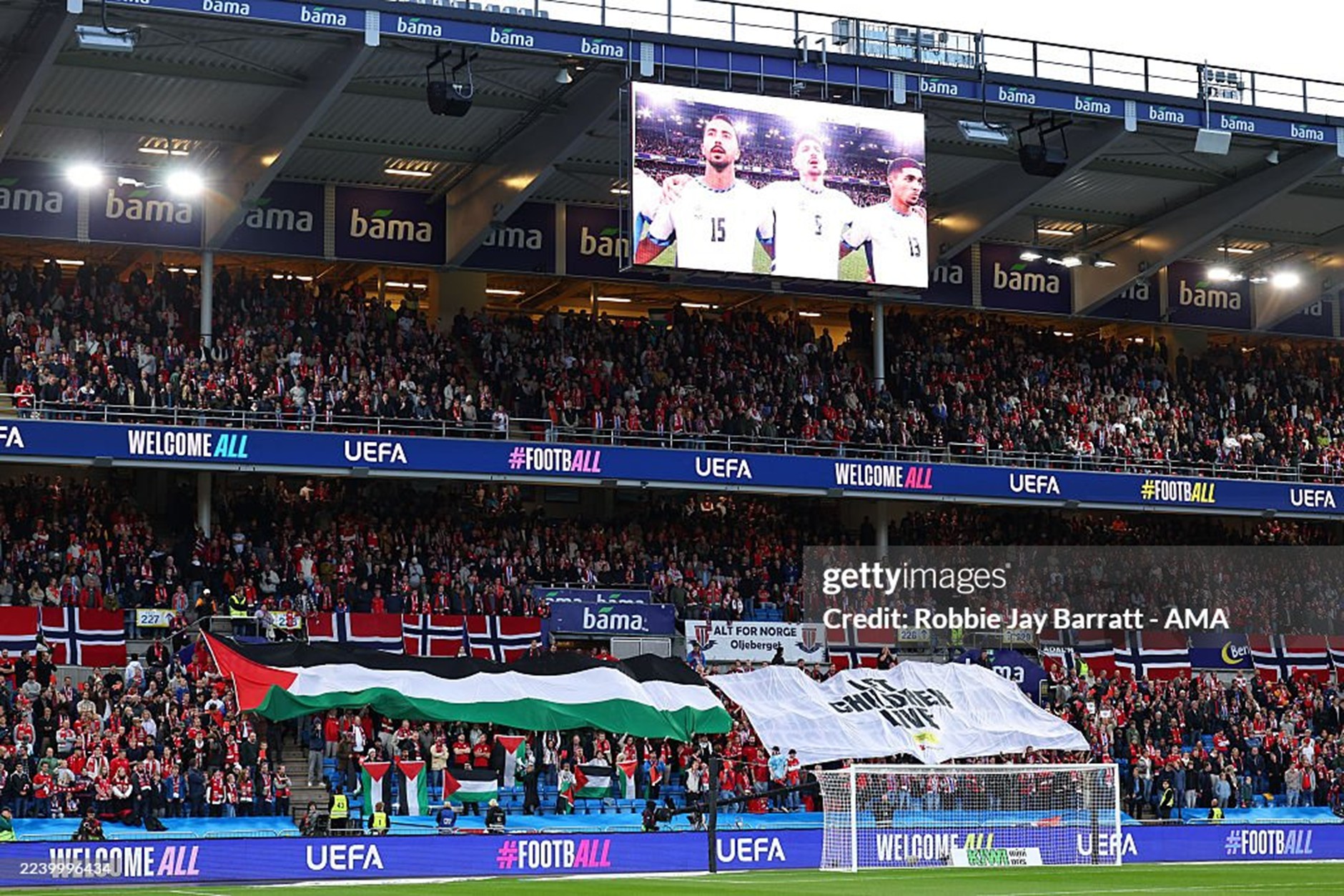 Packed football stadium crowd in red, waving flags under bright stadium lights; banners read 'WELCOME ALL' and 'UEFA'.