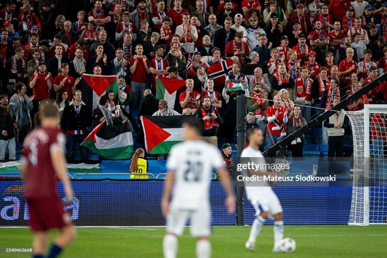 Crowd of soccer fans in red shirts, waving Palestinian flags behind the goal, with players on the field in the foreground.