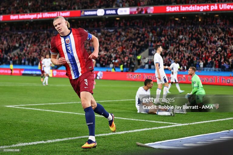 Soccer player in red-and-blue kit sprinting on the pitch near the goal as a crowded stadium watches nearby.