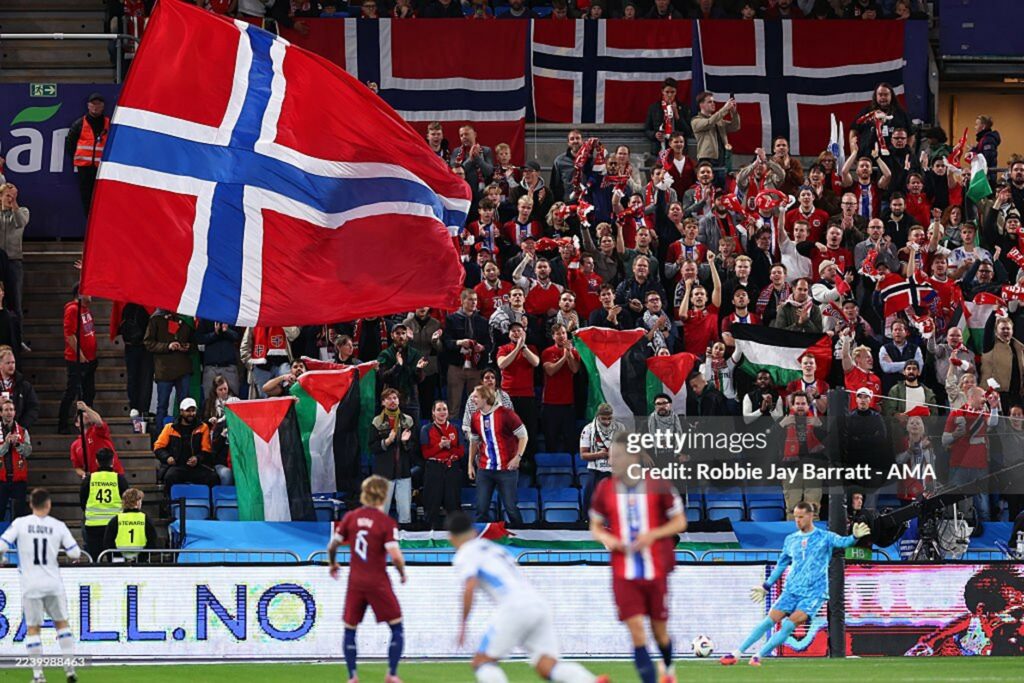 norwegia vs israel 5 dompet dhuafa Packed stadium crowd cheer as fans wave flags, dominated by a large Norwegian flag; Palestinian flags visible among front rows, players on the pitch below.