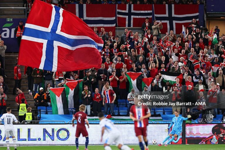 A large Norwegian flag waves over a packed stadium crowd during a match at the stadium stands.