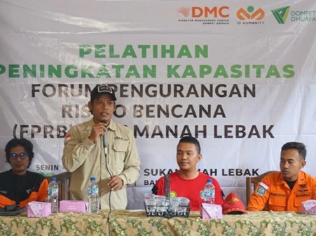 pelatihanbencanadmcdilebak2 dompet dhuafa Man in a beige uniform speaks into a microphone at a disaster risk reduction training panel; banner behind reads 'Pelatihan Peningkatan Kapasitas...' with three other panelists seated at a table.