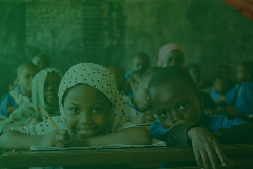 pendidikan anak yatim dompet dhuafa Two smiling students sit at a classroom desk in the foreground—a girl in a dotted hijab writing, beside a boy resting his chin on the desk, with classmates in the background.