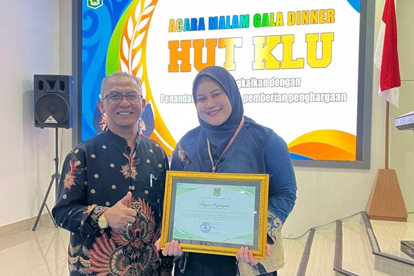 penghargaan untuk bidan untuk negeri 3 dompet dhuafa Two people pose indoors at an award ceremony; man in a batik shirt and glasses stands beside a woman in a blue hijab holding a framed certificate, with a 'HUT KLU' banner in the background.