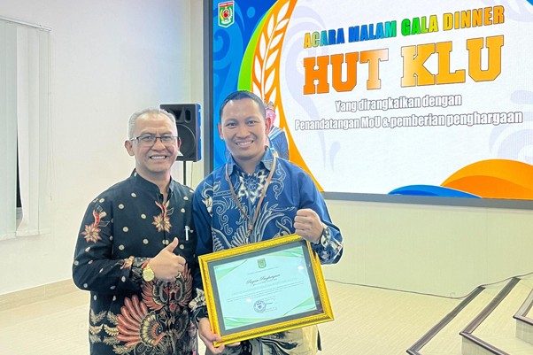 penghargaan untuk bidan untuk negeri 5 dompet dhuafa Two men at an awards ceremony; one hands a framed certificate to the other in front of a colorful banner.