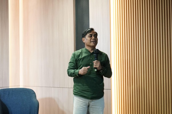 refleksijatidiri11 dompet dhuafa Man in a green shirt giving a talk, holding a microphone in a conference room with wooden wall panels on the side.
