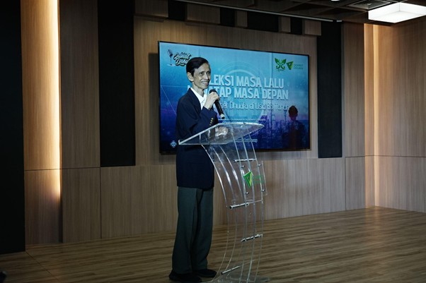 refleksijatidiri6 dompet dhuafa Man in a dark suit speaks at a clear acrylic podium on a wooden stage, with a blue conference screen behind him showing Indonesian text.