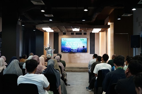 refleksijatidiri7 dompet dhuafa Speaker presents at a podium on a raised stage, addressing a seated audience in a dim conference room with a large screen behind him.