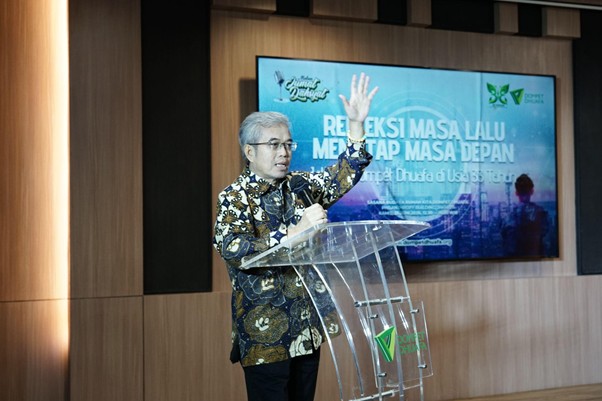 refleksijatidiri8 dompet dhuafa Male presenter in batik shirt speaks at a clear podium, raising one hand as he presents; blue seminar screen behind him shows Indonesian text.