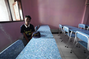 Smiling student in a dark polo sits at a blue speckled desk in a pink-walled classroom.