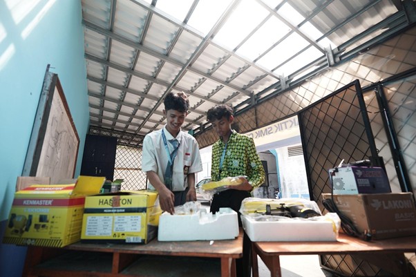 ruang praktikum di smk mitra permata 9 dompet dhuafa Two young men in a workshop examine tools laid out on a table, surrounded by boxes and foam packaging in a bright, open shed with a metal roof.