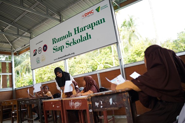rumoh harapan siap sekolah aceh 1 dompet dhuafa Students in brown hijabs at wooden desks in a classroom under a banner reading 'Rumah Harapan Siap Sekolah,' with a teacher assisting them.