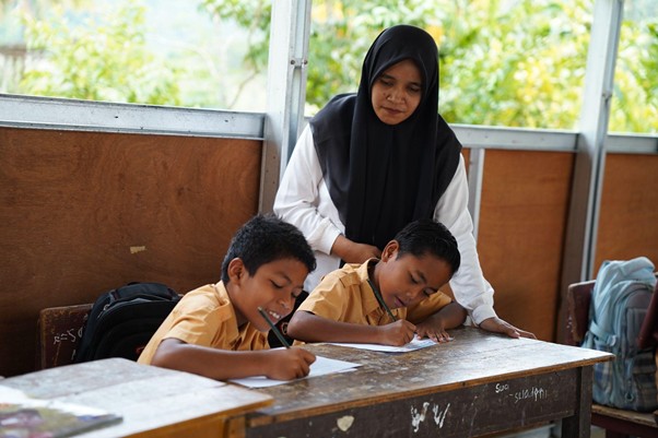 rumoh harapan siap sekolah aceh 10 dompet dhuafa Teacher in a black hijab stands behind two young students who are seated at a wooden desk, helping them as they write on papers in a classroom setting.