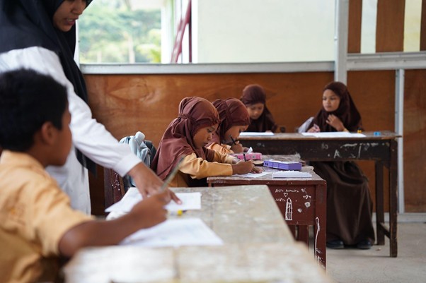 Teacher in hijab helping students as young girls in brown hijabs write at desks in a classroom