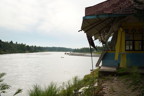 rumoh harapan siap sekolah aceh 2 dompet dhuafa Riverside scene with a colorful wooden shack on the right, grassy bank, and a wide river with small boats in the distance.