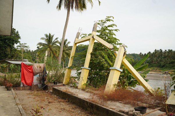 Leaning yellow concrete gate posts with a toppled crossbeam beside a river and tropical plants in the yard.