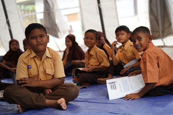 rumoh harapan siap sekolah aceh 4 dompet dhuafa Group of smiling children in tan and brown school uniforms seated on a blue mat inside a tent classroom.