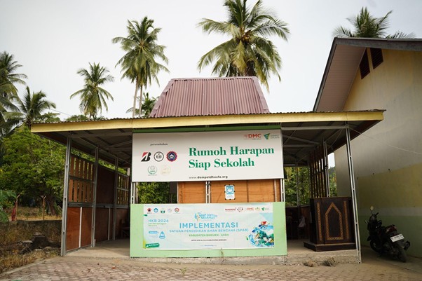 Small open-air community booth with banners reading 'Rumoh Harapan Siap Sekolah' and logos, set among palm trees and a brick-paved area.