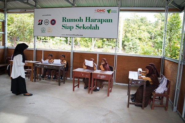 Female teacher stands by students seated at desks in an outdoor classroom; banner reads 'Rumoh Harapan Siap Sekolah'.