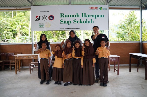 Group of eight Indonesian children in yellow uniforms with brown skirts/pants, two adults behind, under a banner reading Rumoh Harapan Siap Sekolah (school readiness program).