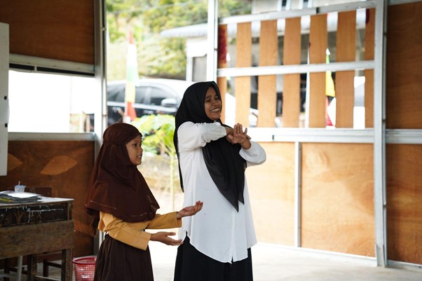 Two girls wearing hijabs practice a coordinated hand gesture indoors, smiling as they move.