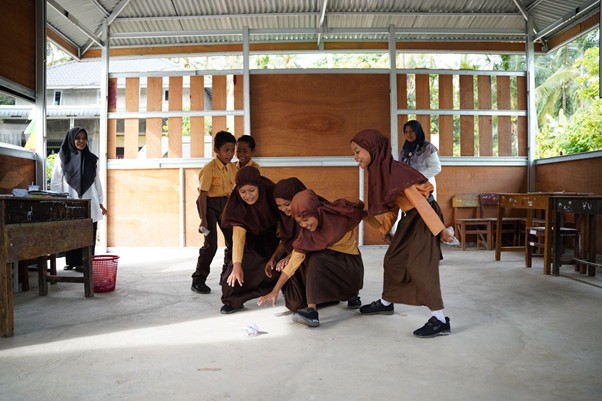 Group of students in brown uniforms in a covered outdoor area, bending to pick up a small paper object on the ground.