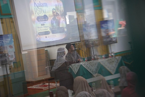 Panel of two speakers seated at a table with microphones; projection screen shows a colorful poster, audience members in headscarves watching the talk.”] ,