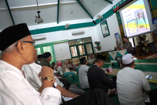 safari dakwah ustaz sugimoto di depok 6 dompet dhuafa Men in traditional caps sit on a green-carpeted floor in a mosque-like hall, listening to a speaker at the front with a projector screen.