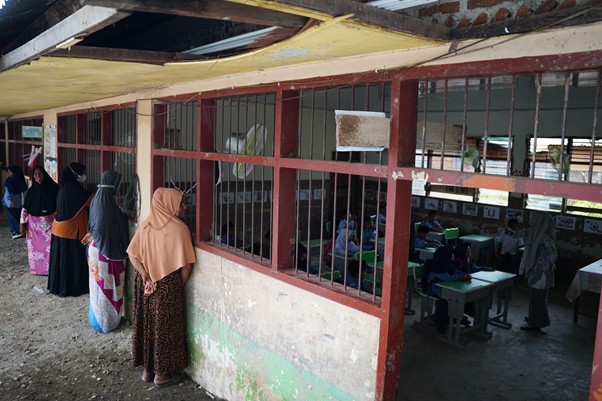 Line of women in hijabs standing outside a classroom with barred windows; students sit at desks inside.