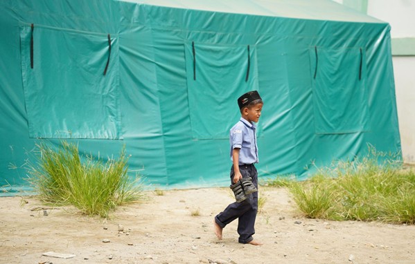 Young boy in a police-style uniform walks barefoot on dirt, carrying a baseball glove beside a teal tent.
