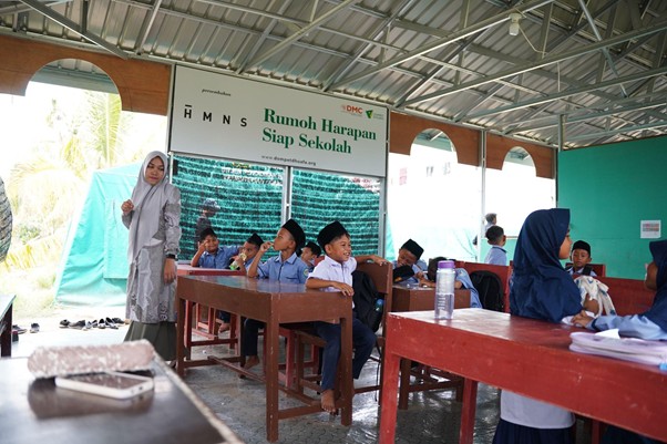 Children in blue uniforms sit at desks under a sheltered outdoor area; a teacher in a gray hijab stands to the left near a banner that reads Rumoh Harapan Siap Sekolah.