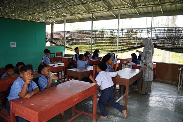 A classroom with young students in blue uniforms seated at wooden desks while a teacher stands at the front giving instruction.