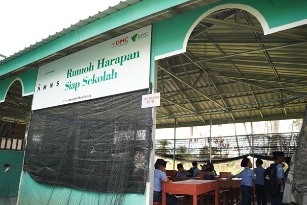 Outdoor school shelter with green trim and a sign reading 'Rumah Harapan Siap Sekolah'; students in blue uniforms sit at desks under a metal roof.