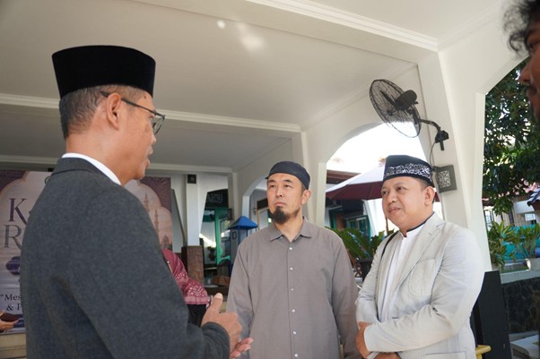 Three men in traditional Muslim attire converse on a covered porch, one facing away while two look on thoughtfully.