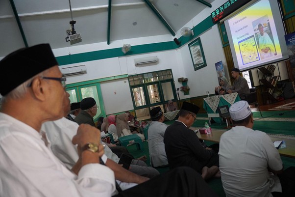 Men in traditional clothing sit on a green-carpeted floor in a mosque, listening to a speaker presenting slides at the front.