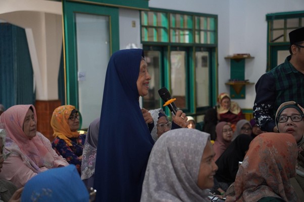 Woman in a blue hijab speaks into a handheld microphone at a crowded community gathering.