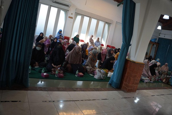 Groups of women in hijabs seated on green mats in a bright room during a community gathering, with bags and bottles nearby.